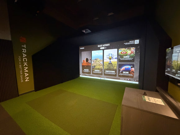 Trackman indoor golf simulator room with a large projection screen showing game options, green turf hitting surface, and a wooden control console in a dark enclosed space.
